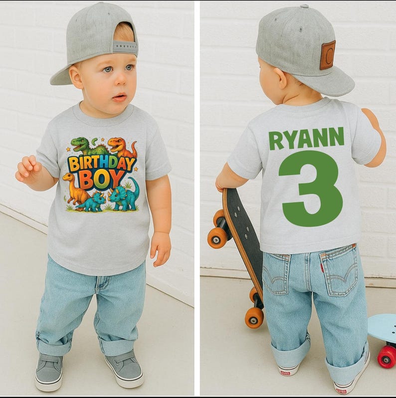 May include: A light gray t-shirt with a colorful dinosaur graphic and the text "BIRTHDAY BOY." The back of the shirt has the name "RYANN" and the number "3" in green. The child is wearing light blue jeans and a gray baseball cap.