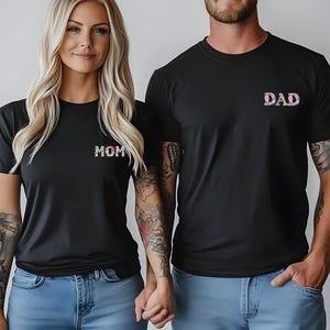 May include: Two black short-sleeved crew neck t-shirts. The left shirt features the word MOM in a floral pattern on the chest. The right shirt features the word DAD in a floral pattern on the chest. Both shirts are worn with blue denim jeans.