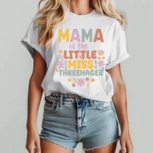 May include: A white short-sleeve t-shirt featuring the colorful text MAMA of the LITTLE MISS THREENAGER in yellow, orange, pink, and teal. The design is decorated with small floral illustrations in matching pastel colors around the lettering.