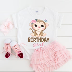 May include: White t-shirt with a brown monkey wearing a pink flower crown and a pink butterfly above it. The text "Birthday girl" is printed below the monkey in brown and pink letters.