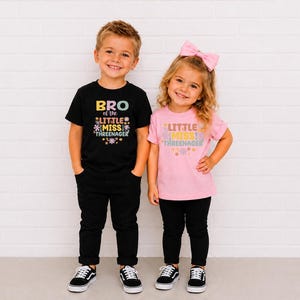 May include: A black t-shirt with the text BRO OF THE LITTLE MISS THREENAGER and a pink t-shirt with the text LITTLE MISS THREENAGER, both featuring colorful floral graphics. The shirts are paired with black pants and black sneakers.