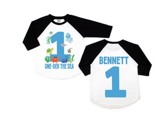May include: A white and black baseball style shirt with a blue number one and the text "ONE-DER THE SEA" on the front. The back of the shirt has the name "BENNETT" and a blue number one.
