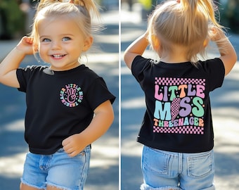 May include: A black t-shirt with a rainbow graphic that says "Little Miss Three-nager" on the back and a rainbow graphic that says "Three-nager Little Miss" on the front. The shirt is being worn by a child with blonde hair.