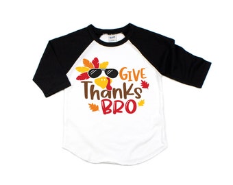 May include: A white and black raglan-style shirt with a Thanksgiving graphic. The design features a turkey wearing sunglasses and the words "Give Thanks Bro". The shirt has black sleeves and is suitable for children.