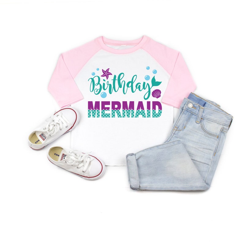 May include: White and pink raglan t-shirt with the text "Birthday Mermaid" in teal and purple with a mermaid tail, a seashell, and a starfish. The shirt is laying on a white surface with a pair of white sneakers and light blue jeans.
