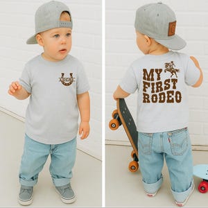 Op de afbeelding: Lichtgrijs t-shirt met een hoefijzer en de naam "LUCAS" in bruin. Op de achterkant staat "MY FIRST RODEO" met een cowboy-afbeelding. Het kind draagt een lichtblauwe spijkerbroek en een grijze baseballcap. Het kind houdt een skateboard vast.