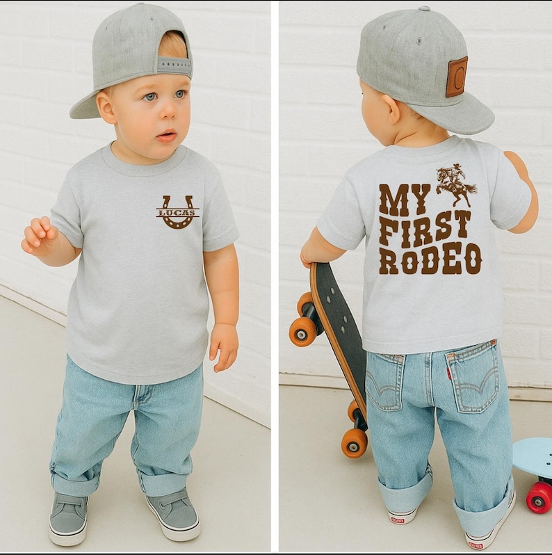 Op de afbeelding: Lichtgrijs t-shirt met een hoefijzer en de naam "LUCAS" in bruin. Op de achterkant staat "MY FIRST RODEO" met een cowboy-afbeelding. Het kind draagt een lichtblauwe spijkerbroek en een grijze baseballcap. Het kind houdt een skateboard vast.