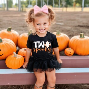 Pu&ograve; includere: Una bambina indossa una maglietta nera con la scritta "TWO spooky" e una grafica di un fantasma, abbinata a un tut&ugrave; nero e un fiocco rosa. Lo sfondo presenta diverse zucche arancioni, suggerendo un tema di Halloween.