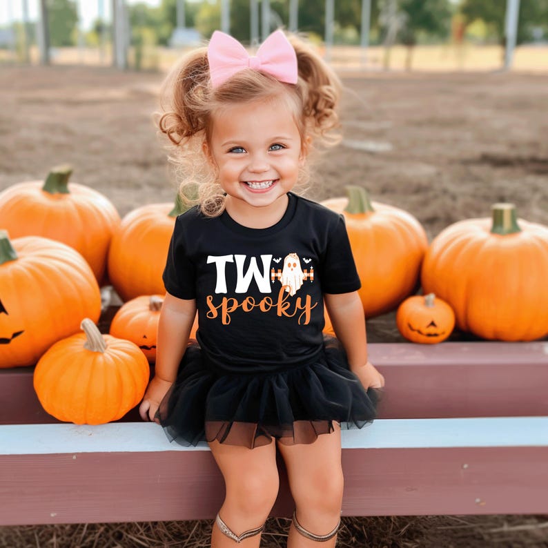 Pu&ograve; includere: Una bambina indossa una maglietta nera con la scritta "TWO spooky" e una grafica di un fantasma, abbinata a un tut&ugrave; nero e un fiocco rosa. Lo sfondo presenta diverse zucche arancioni, suggerendo un tema di Halloween.