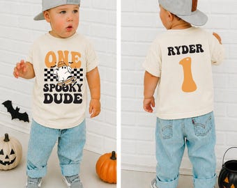 May include: A cream-colored t-shirt featuring a graphic of a ghost on a skateboard with the text ONE SPOOKY DUDE on the front and the name RYDER with the number 1 on the back, paired with light blue denim jeans and a gray baseball cap.