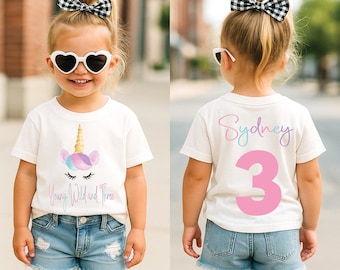 May include: A white t-shirt with a unicorn graphic and the text "Young Wild and Three". The back of the shirt has the name "Sydney" and the number "3" in pink. The child is wearing denim shorts and heart-shaped sunglasses.