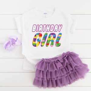 May include: White t-shirt with the text "Birthday Girl" in pink letters with a colourful Lego block design. The shirt is paired with a purple tulle skirt.