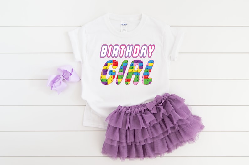May include: White t-shirt with the text "Birthday Girl" in pink letters with a colourful Lego block design. The shirt is paired with a purple tulle skirt.