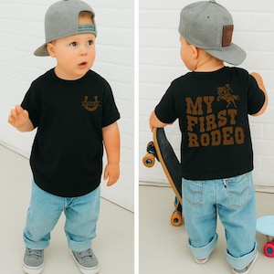 Op de afbeelding: Een zwart t-shirt met de tekst "LUCKY" in een hoefijzerontwerp. De achterkant van het shirt heeft de tekst "MY FIRST RODEO" met een afbeelding van een cowboy op een paard. Het kind draagt een grijze baseballcap en lichtblauwe jeans.