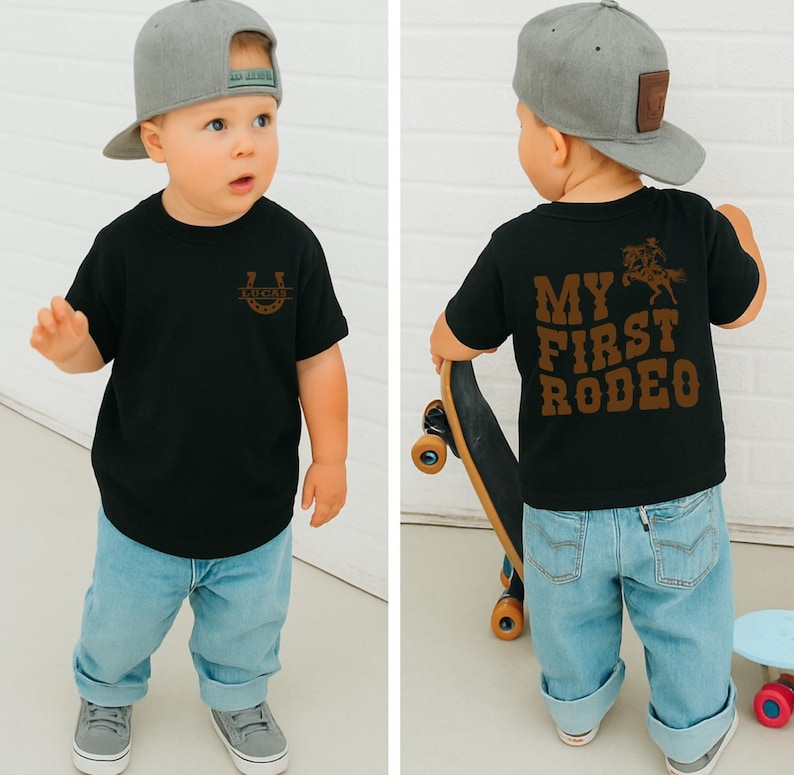 Op de afbeelding: Een zwart t-shirt met de tekst "LUCKY" in een hoefijzerontwerp. De achterkant van het shirt heeft de tekst "MY FIRST RODEO" met een afbeelding van een cowboy op een paard. Het kind draagt een grijze baseballcap en lichtblauwe jeans.