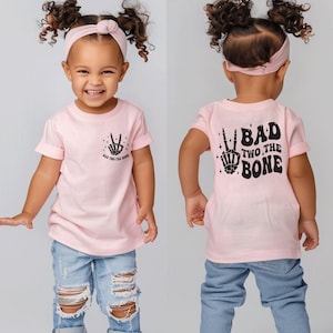 Puede incluir: Camiseta rosa para ni&ntilde;o peque&ntilde;o con una mano de esqueleto negra haciendo un signo de paz y el texto "Bad Two The Bone".