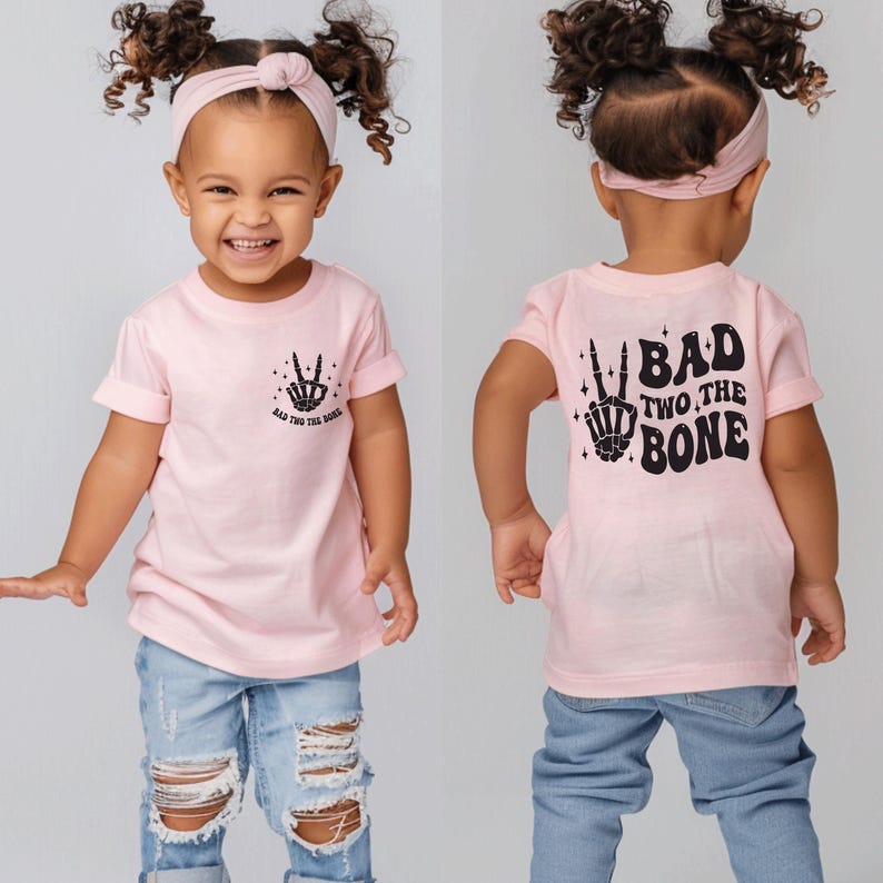 Puede incluir: Camiseta rosa para ni&ntilde;o peque&ntilde;o con una mano de esqueleto negra haciendo un signo de paz y el texto "Bad Two The Bone".