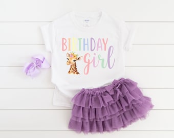 Pode incluir: Camiseta branca com as palavras "Birthday Girl" em cores pastel e um gráfico de girafa. O conjunto inclui uma saia tutu roxa e um laço a condizer. Perfeito para uma festa de aniversário.