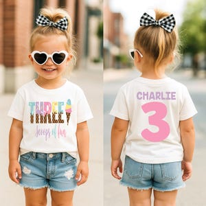 May include: A white t-shirt with the text "THREE" and "scoops of fun" in colorful lettering, with ice cream cone graphics. The back of the shirt has the name "CHARLIE" and the number "3" in pink. The child is wearing denim shorts and heart-shaped sunglasses.
