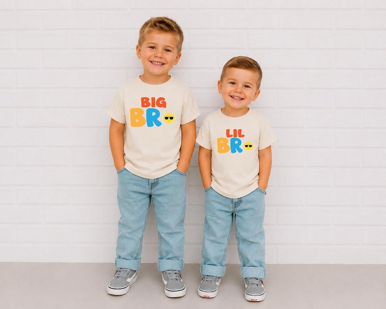 May include: Two cream-coloured short-sleeve t-shirts featuring colourful graphic text. One shirt displays the text BIG BRO in red, orange, and blue with a yellow smiley face wearing sunglasses. The other shirt displays LIL BRO in the same style.