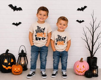 May include: Two beige short-sleeved t-shirts featuring black and orange text. One shirt reads "the spooky ONE'S BRO" with a small bat and spider graphic. The other shirt reads "the spooky ONE" with a small bat and spider graphic.