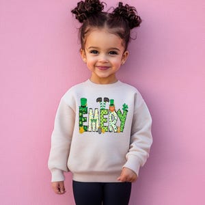 May include: A child wearing a cream colored sweatshirt with a green, gold, and orange design that says "EMERY".
