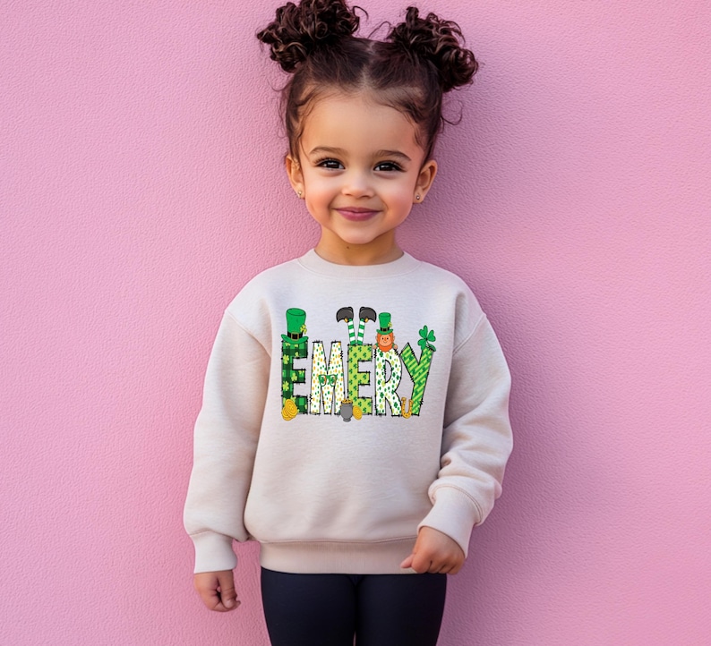 May include: A child wearing a cream colored sweatshirt with a green, gold, and orange design that says "EMERY".