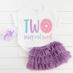 May include: White t-shirt with the text "TWO sassy and sweet" and a pink donut with sprinkles. The shirt is paired with a purple tulle skirt.