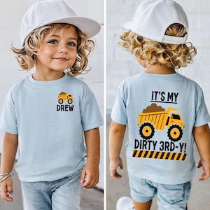 May include: A light blue short-sleeve t-shirt featuring a small yellow dump truck graphic and the name DREW on the front. The back displays a large yellow dump truck graphic with the text IT'S MY DIRTY 3RD-Y! above a yellow and black striped border.