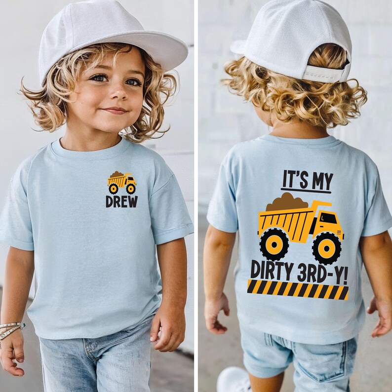 May include: A light blue short-sleeve t-shirt featuring a small yellow dump truck graphic and the name DREW on the front. The back displays a large yellow dump truck graphic with the text IT'S MY DIRTY 3RD-Y! above a yellow and black striped border.
