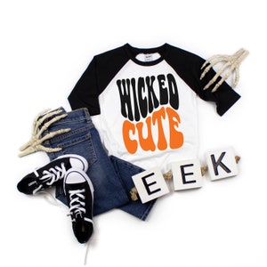 May include: A white and black raglan shirt with the words "WICKED CUTE" in black and orange. The shirt is paired with blue jeans and black sneakers. Decorative skeleton hands and "EEK" letters complete the Halloween-themed ensemble.