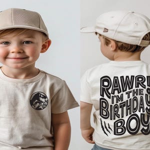 May include: A toddler wearing a beige t-shirt with a dinosaur graphic on the front and the text "RAWR! I'M THE BIRTHDAY BOY" on the back. The toddler is also wearing a beige baseball cap.