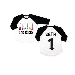 May include: White and black baseball style shirt with a graphic of six colorful guitars and the text "One Rocks". The back of the shirt has the name "Seth" and the number "1" in black.