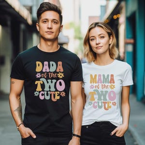 May include: A black t-shirt with the text DADA of the TWO CUTE and a white t-shirt with the text MAMA of the TWO CUTE. Both shirts feature colorful, retro-style lettering in shades of pink, yellow, blue, and orange, accented with small flower icons.