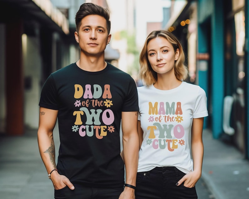 May include: A black t-shirt with the text DADA of the TWO CUTE and a white t-shirt with the text MAMA of the TWO CUTE. Both shirts feature colorful, retro-style lettering in shades of pink, yellow, blue, and orange, accented with small flower icons.