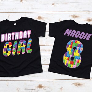 May include: Two black t-shirts with a birthday theme. One shirt says "Birthday Girl" in pink letters with a colourful Lego design. The other shirt has the name "Maddie" in pink letters and a large number "8" made of colourful Lego blocks.