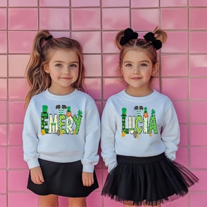 May include: Two girls wearing white sweatshirts with green shamrock designs and the names "Emery" and "Lucia" on them. The girls are standing in front of a pink tiled wall.