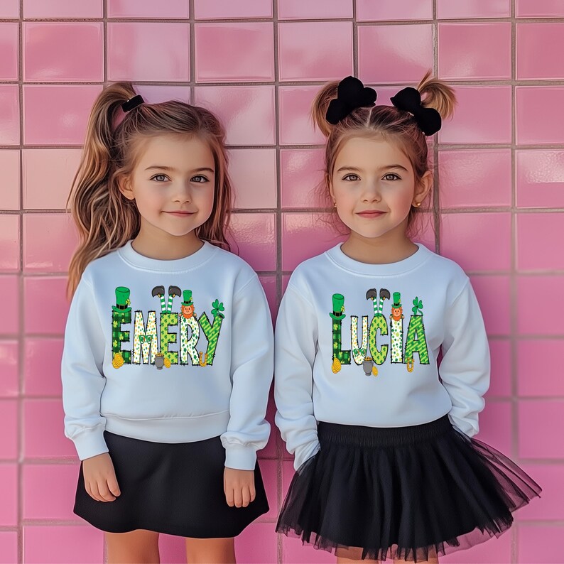 May include: Two girls wearing white sweatshirts with green shamrock designs and the names "Emery" and "Lucia" on them. The girls are standing in front of a pink tiled wall.
