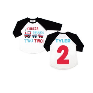 May include: A white and black baseball style shirt with a red and blue train design on the front. The text "CHUGGA CHUGGA TWO TWO!" is printed on the shirt. The back of the shirt has the name "TYLER" and the number "2" printed in red.
