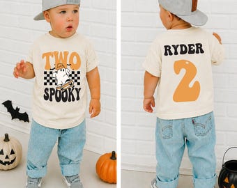 May include: A cream-colored t-shirt featuring a graphic of a ghost on a skateboard with the text "TWO SPOOKY" on the front and the name "RYDER" with the number "2" on the back, paired with light blue denim jeans and a gray baseball cap.