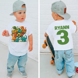 May include: A toddler wearing a white t-shirt with a colorful dinosaur graphic and the text "BIRTHDAY BOY". The child is also wearing blue jeans and a gray baseball cap. The back of the shirt has the name "RYANN" and the number "3".