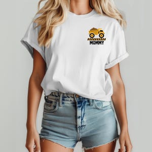 May include: A white short-sleeve t-shirt featuring a small graphic on the left chest. The graphic depicts a yellow dump truck above black and yellow caution stripes and the word MOMMY printed in bold black capital letters.
