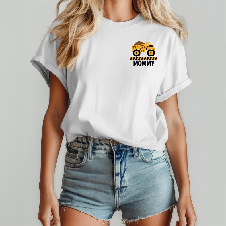 May include: A white short-sleeve t-shirt featuring a small graphic on the left chest. The graphic depicts a yellow dump truck above black and yellow caution stripes and the word MOMMY printed in bold black capital letters.