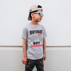 Puede incluir: Camiseta gris con las palabras "BIRTHDAY BOY" y un gr&aacute;fico de motocicleta. La camiseta tiene una franja a cuadros rojos y blancos en la parte inferior. La persona lleva una gorra de b&eacute;isbol beige y gafas de sol azules.