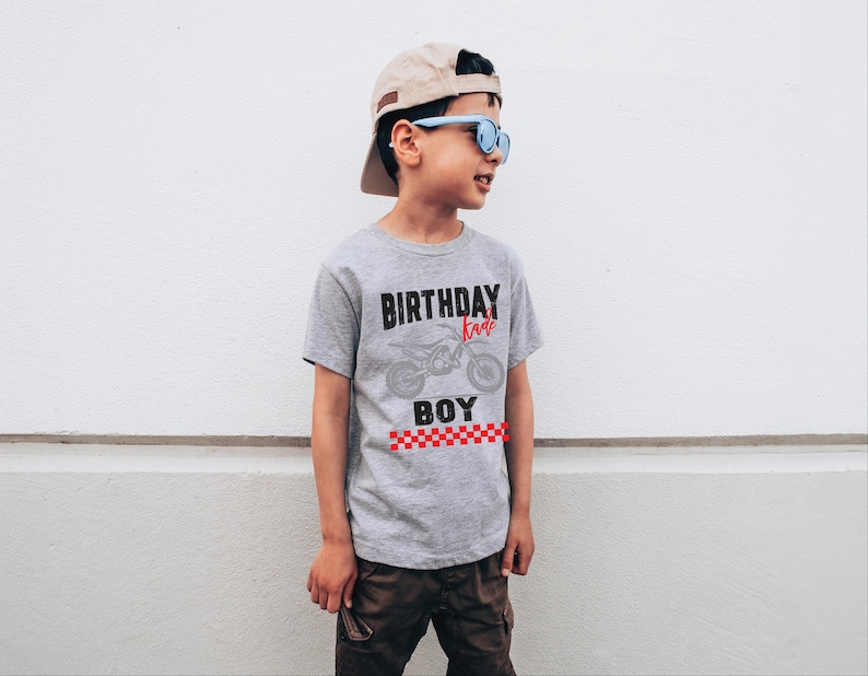 Puede incluir: Camiseta gris con las palabras "BIRTHDAY BOY" y un gr&aacute;fico de motocicleta. La camiseta tiene una franja a cuadros rojos y blancos en la parte inferior. La persona lleva una gorra de b&eacute;isbol beige y gafas de sol azules.