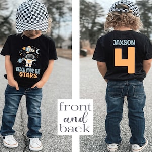 May include: Black t-shirt with a graphic of an astronaut and the text "Reach for the Stars". The back of the shirt has the name "Jaxson" and the number "4" in orange. The shirt is being worn by a child wearing a black and white checkered hat and blue jeans.