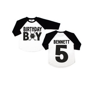 May include: Black and white baseball jersey with the text "Birthday Boy" and a hockey stick and puck graphic on the front. The back of the jersey has the name "BENNETT" and the number "5" in black.