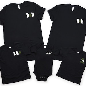 Puede incluir: Camisetas negras y un body negro con texto blanco y gr&aacute;ficos de &aacute;rboles verdes. Las camisetas tienen el texto "DAD", "MOM", "BR" y "CAMP HAPPY CAMPER". El body tiene el texto "SASSY".