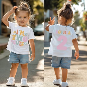 May include: A white t-shirt with the text "I'm Two Cute" in colorful letters and flowers. The back of the shirt has the name "Sophia" in colorful letters and the number "2" in pink. The shirt is being worn by a child.