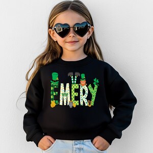 May include: Black sweatshirt with a green, white, and gold design of leprechauns and shamrocks spelling out the name "EMERY".